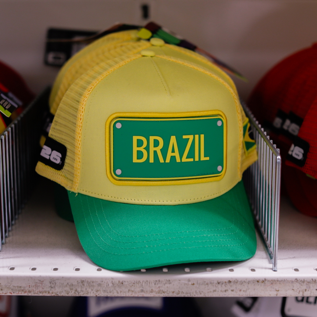 Gul och grön keps med texten "BRAZIL" på framsidan, placerad på en hylla bland andra kepsar.