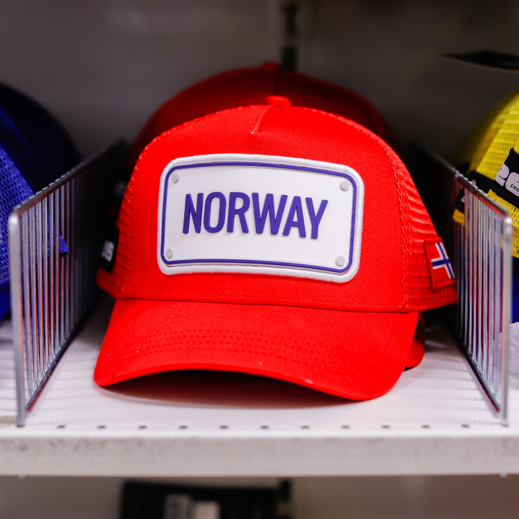 Röd keps med texten "NORWAY" och den norska flaggan, placerad på en hylla bland andra kepsar.