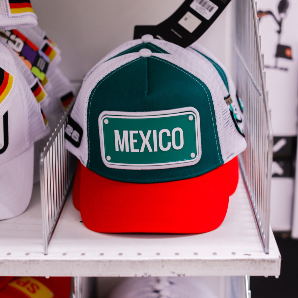 Grön och röd keps med texten "MEXICO" på framsidan, placerad på en hylla bland andra kepsar.