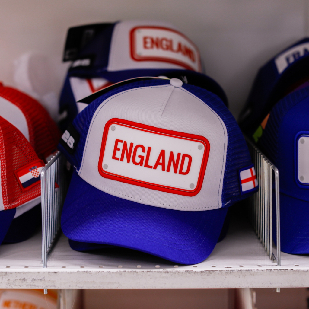 Blå keps med vit och röd etikett som har texten "ENGLAND", placerad bland andra kepsar i en butik.