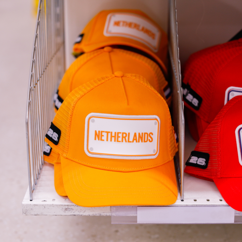 Orange kepsar med texten "NETHERLANDS" i en butik, staplade på en hylla.