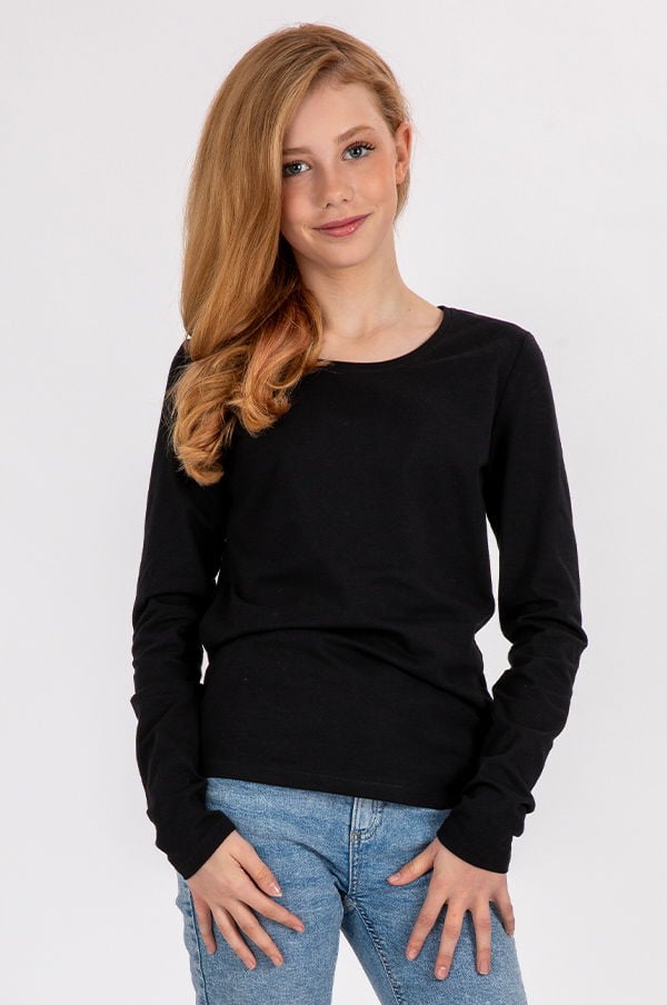 Ung tjej med långt blont hår som bär en svart långärmad tröja och jeans. Hon står med händerna i midjan och ler mot kameran. Bakgrunden är ljus och neutral. Perfekt för mode- och klädesrelaterade sammanhang.