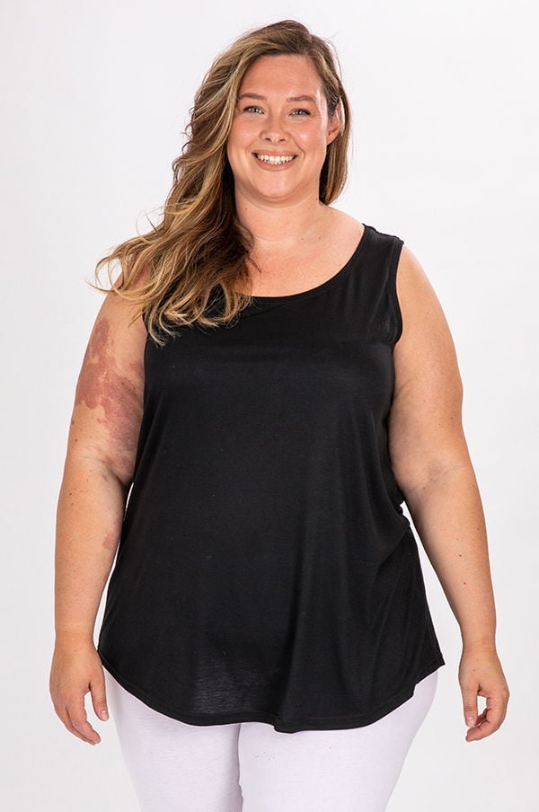 Kvinna med långt hår som ler, iklädd en svart linne och vita byxor, poserar framför en vit bakgrund. Bilden visar en plus size-modell som utstrålar självförtroende och stil.