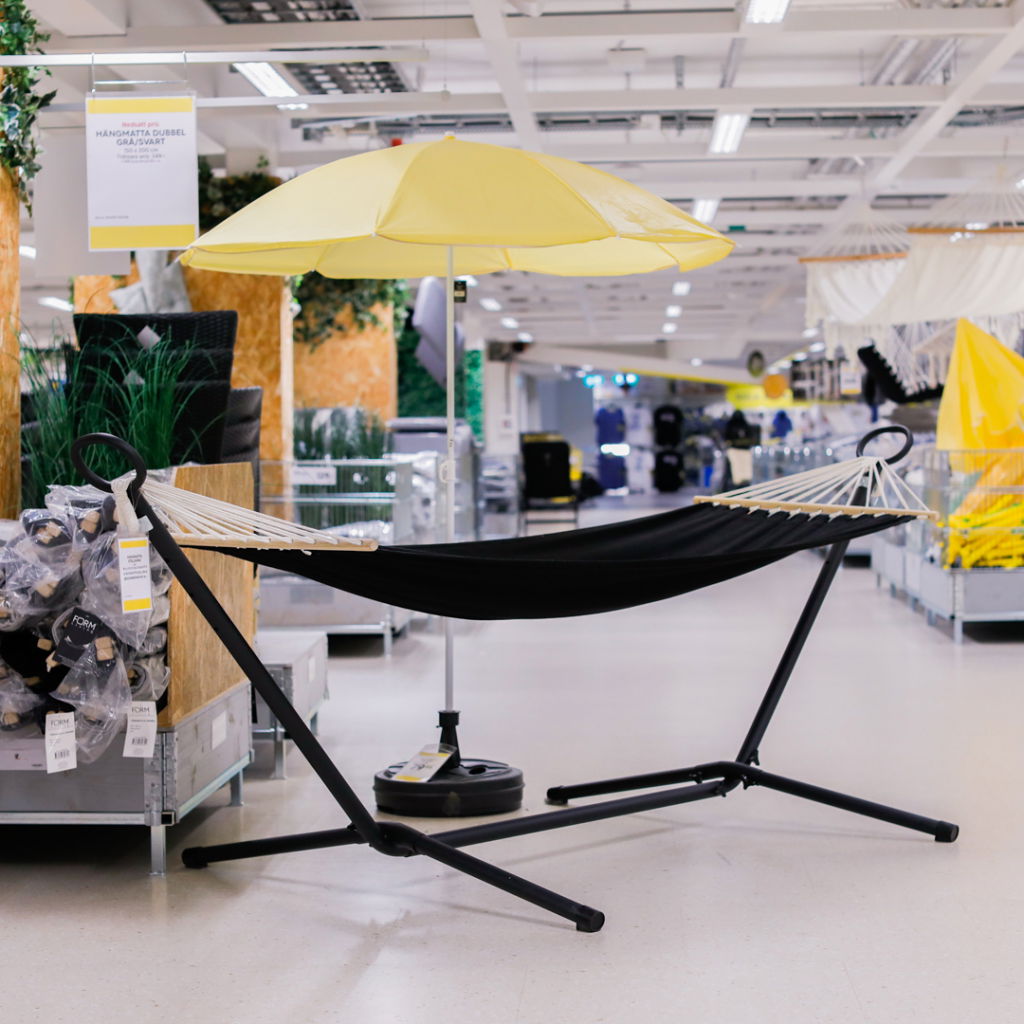 Svart hängmatta med stöd och gul parasoll i en heminredningsbutik, omgiven av gröna växter och inredningsprodukter.