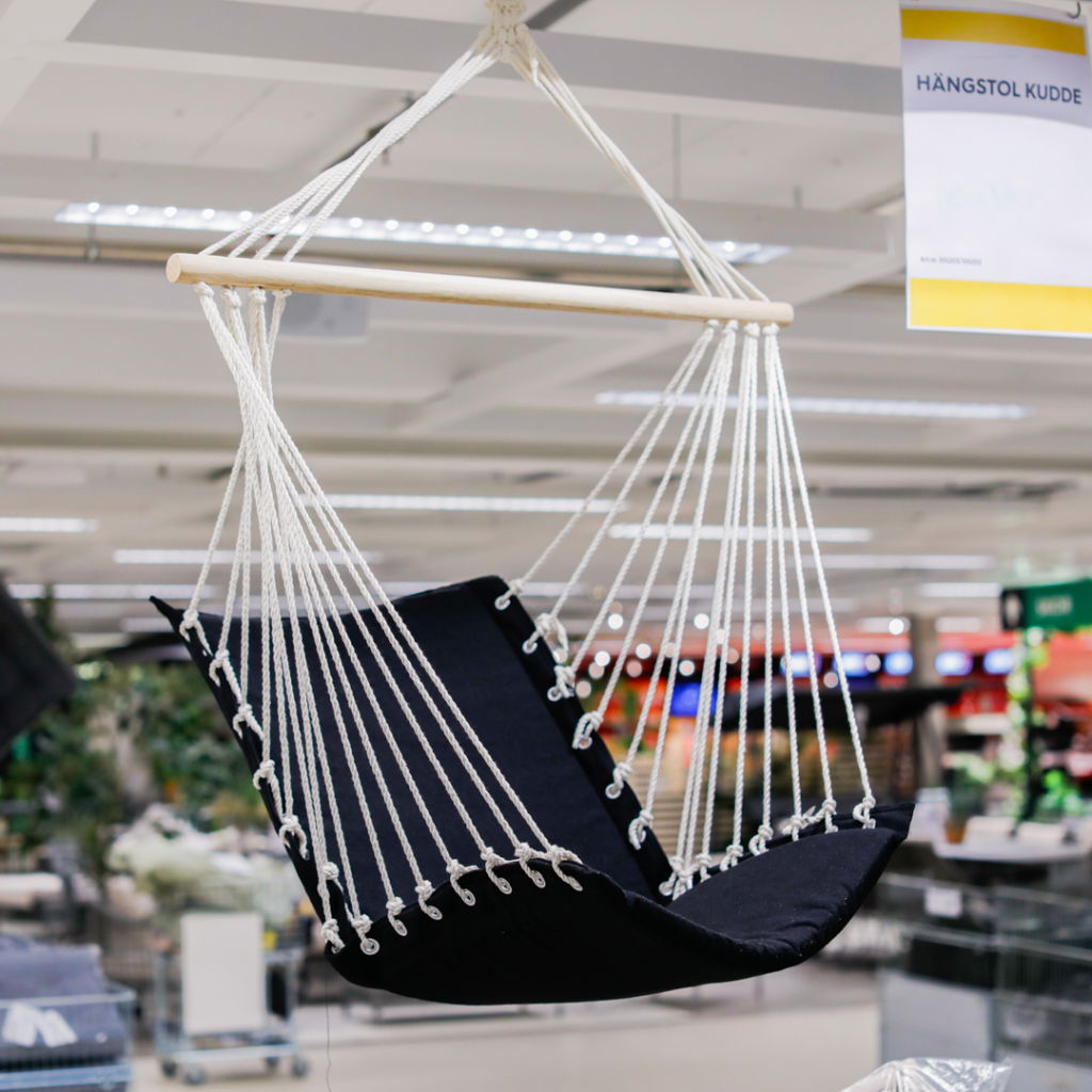 Hängstol i svart tyg med vita rep, hängande från en trästång i en butiksmiljö. Perfekt för avkoppling inomhus eller utomhus.
