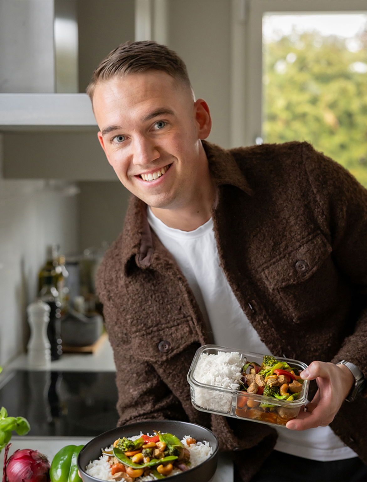 En leende man i köket håller i en skål med ris och grönsaker samt en matlåda med ris och grönsaksrätt. Bakgrunden visar en ljus och modern köksmiljö med grönsaker och oljor synliga.