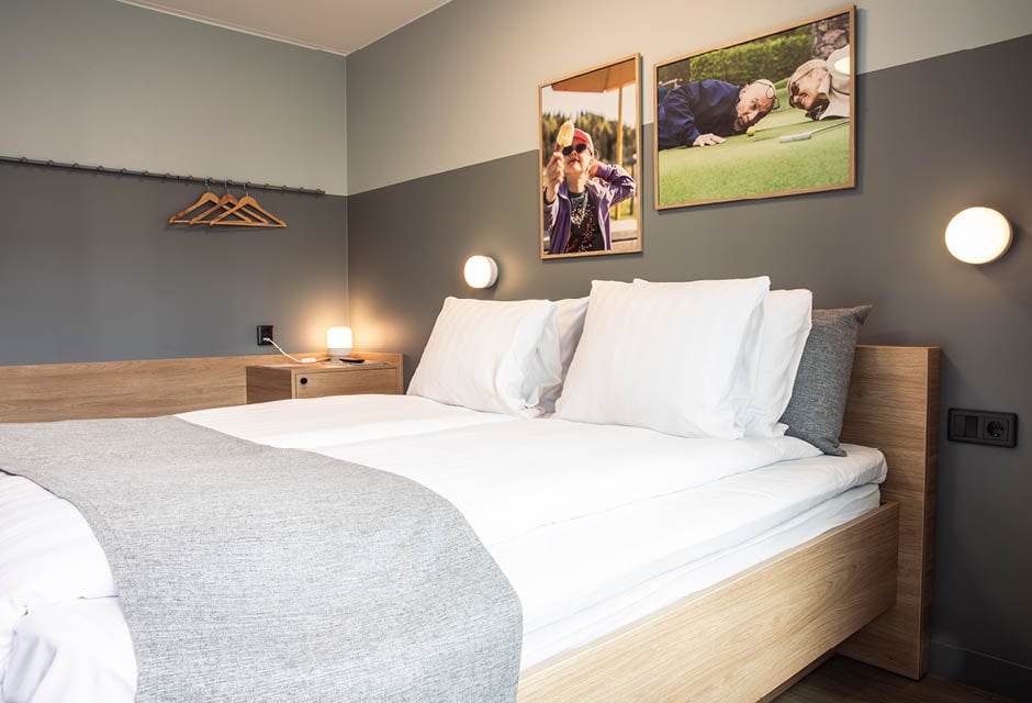Modernt hotellrum med dubbelsäng, ljust trä och grå väggar, prytt med två ramade fotografier. Rummet har en nattlampa, en liten skrivbordsyta och klädhängare, vilket skapar en bekväm och stilren atmosfär.