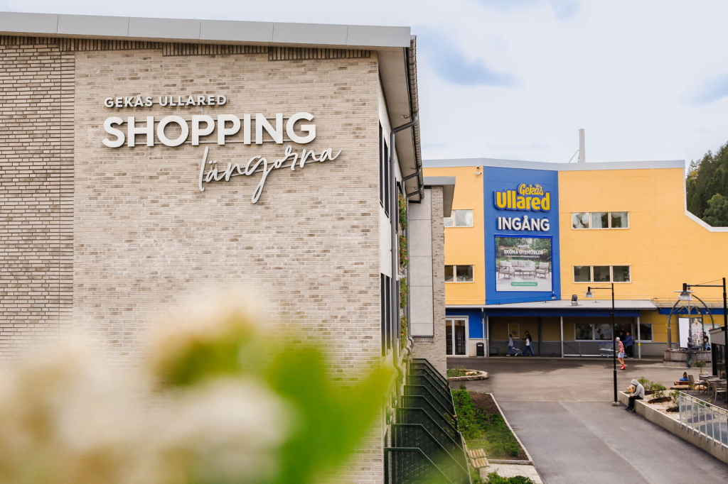 Gekås Ullared shoppingcenter med ingångsskyltar och byggnadens fasad, omgiven av grönska och besökare.