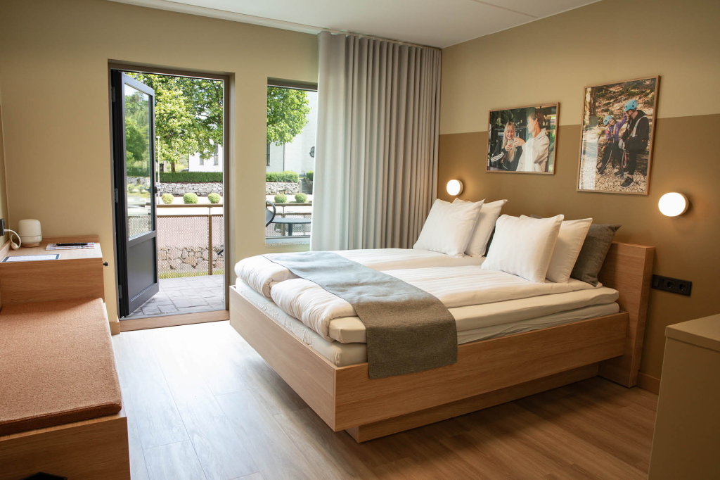 Modernt hotellrum med dubbelsäng, stora fönster och utgång till en balkong. Rummet har en mysig sittgrupp och väggkonst som skapar en inbjudande atmosfär.