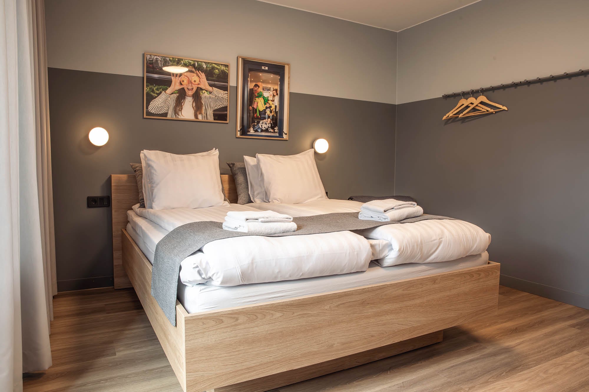 Modern och stilren hotellrum med dubbelsäng, kuddar och handdukar. Väggarna är i gråa toner och rummet har konstverk som dekoration. Det finns även en klädhängare och mjukt golv. Perfekt för en avkopplande vistelse.