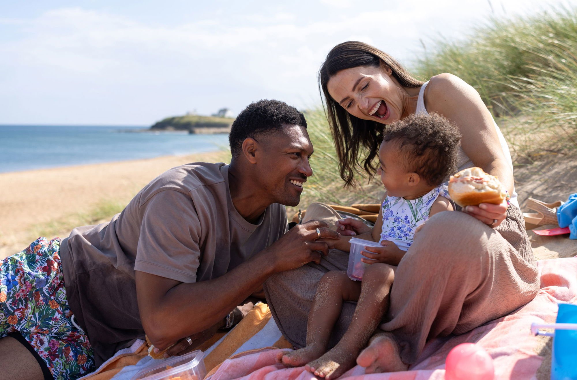 Familj som njuter av en picknick på stranden, leende och lekfulla, med havet i bakgrunden och en solig dag. Barnet sitter på en filt och interagerar glatt med sina föräldrar.