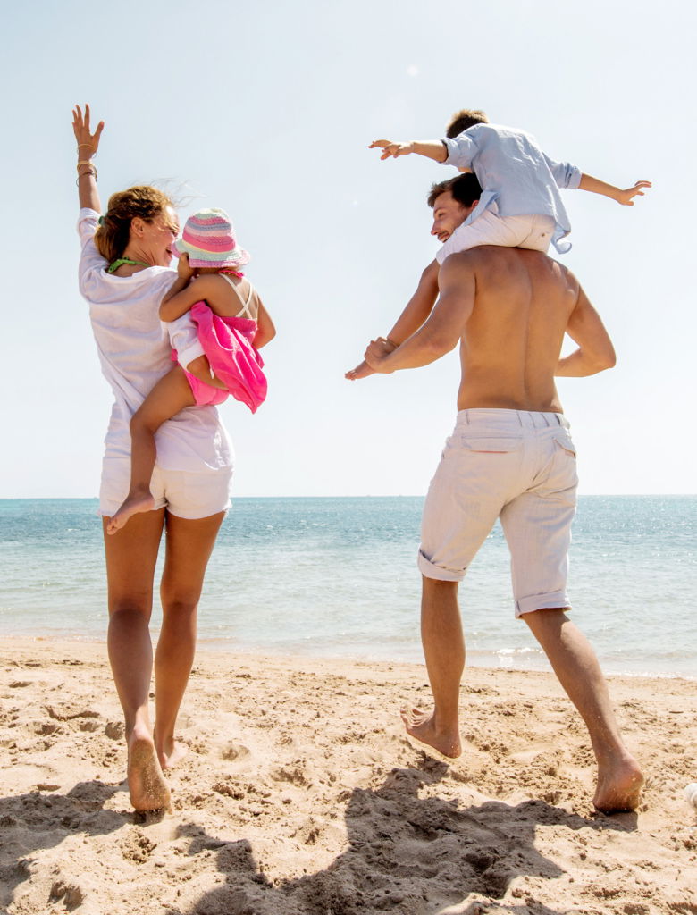 Familj som njuter av en solig dag på stranden, där en mamma bär ett barn och en pappa har en annan på sina axlar, med havet i bakgrunden.