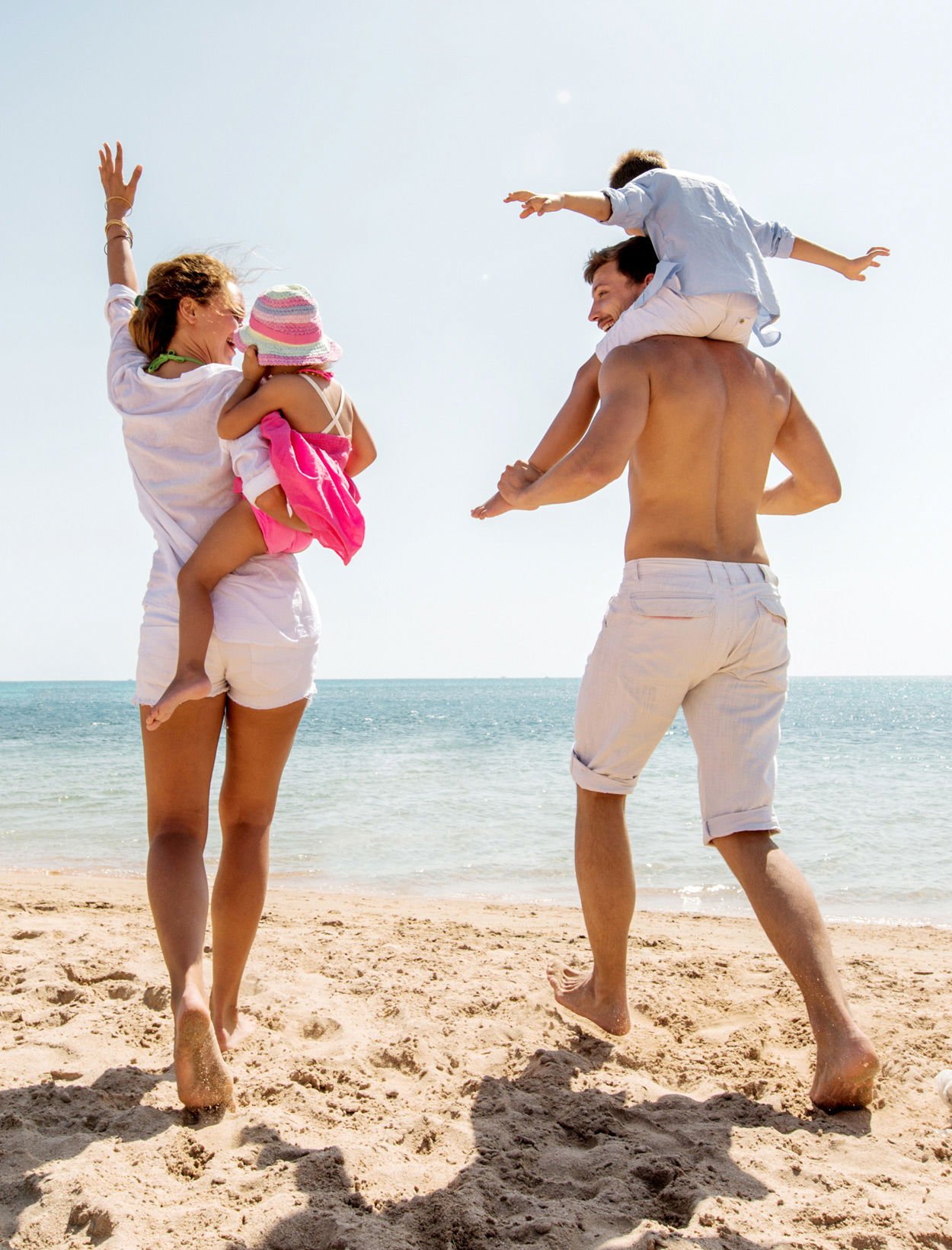 Familj som njuter av en solig dag på stranden, där en mamma bär ett barn och en pappa har en annan på sina axlar, med havet i bakgrunden.