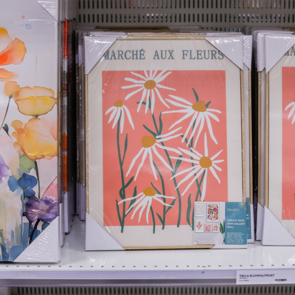 Färgglad tavla med vita prästkragar på korallfärgad bakgrund, förpackad i plast. Tavlan är en del av en utställning med blommotiv och finns på hyllan i en butik.