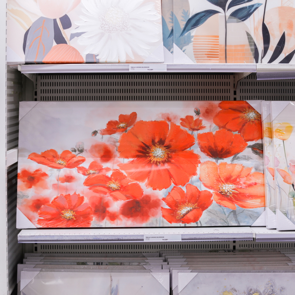 Färgglad tavla med röda blommor, placerad på en hylla bland andra konstverk i en butik.