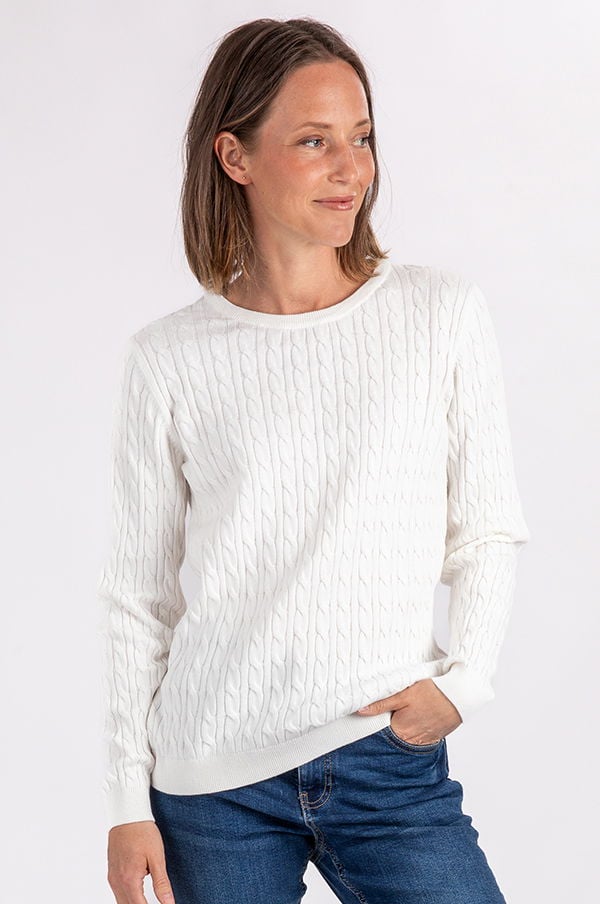 Kvinna i vit kabelstickad tröja, poserar med ett leende. Tröjan har rund halsringning och är kombinerad med blå jeans, vilket ger en avslappnad och stilren look. Idealisk för både vardag och fritid.