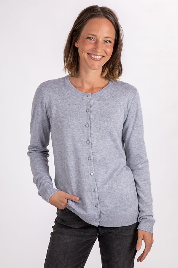 Kvinna i grå cardigan med knappar, leende mot kameran. Hon står med ena handen i fickan och bär svarta jeans. Idealisk bild för mode och klädesbutiker.