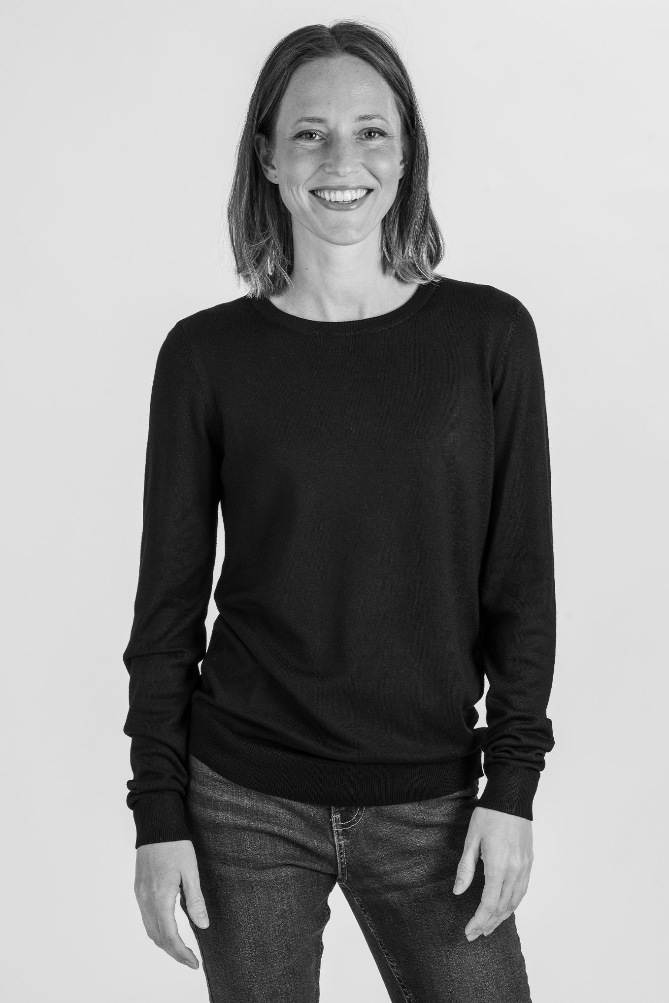 Kvinna med leende, klädd i svart tröja och jeans, poserar framför en ljus bakgrund.