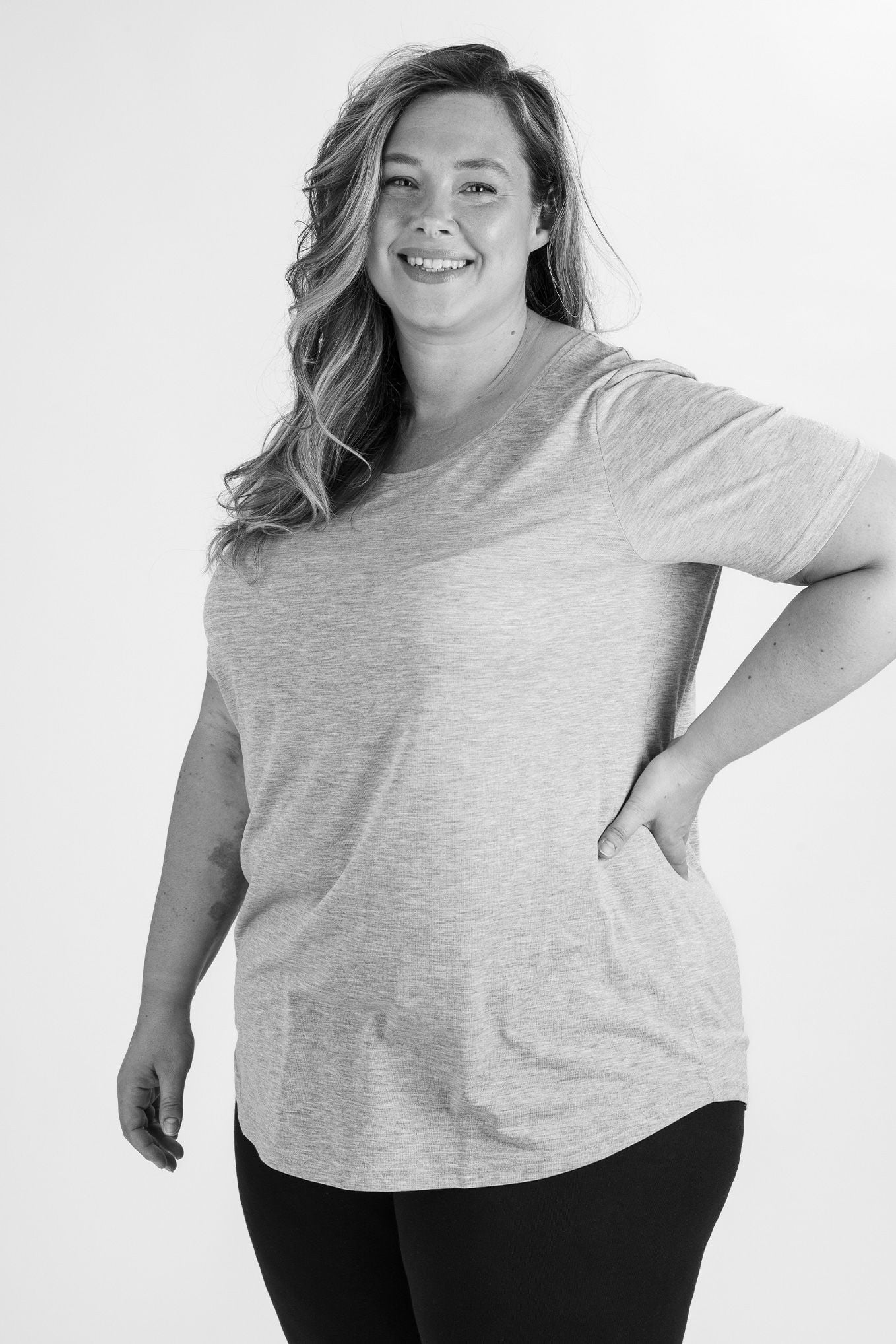 Kvinna med långt hår och leende, klädd i en grå t-shirt och svarta leggings, poserar med en hand på höften. Bilden är i svartvitt och visar en avslappnad och självsäker attityd.
