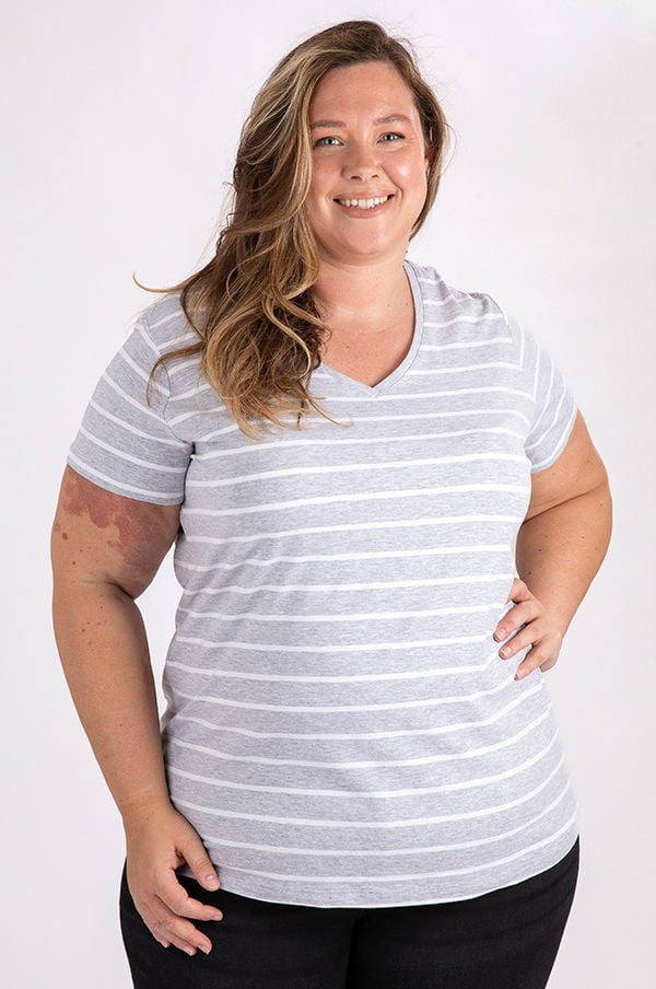 Kvinna med långt hår som ler, iförd en grå randig T-shirt och svarta byxor, står framför en vit bakgrund. T-shirten har en V-ringning och är designad för komfort och stil.