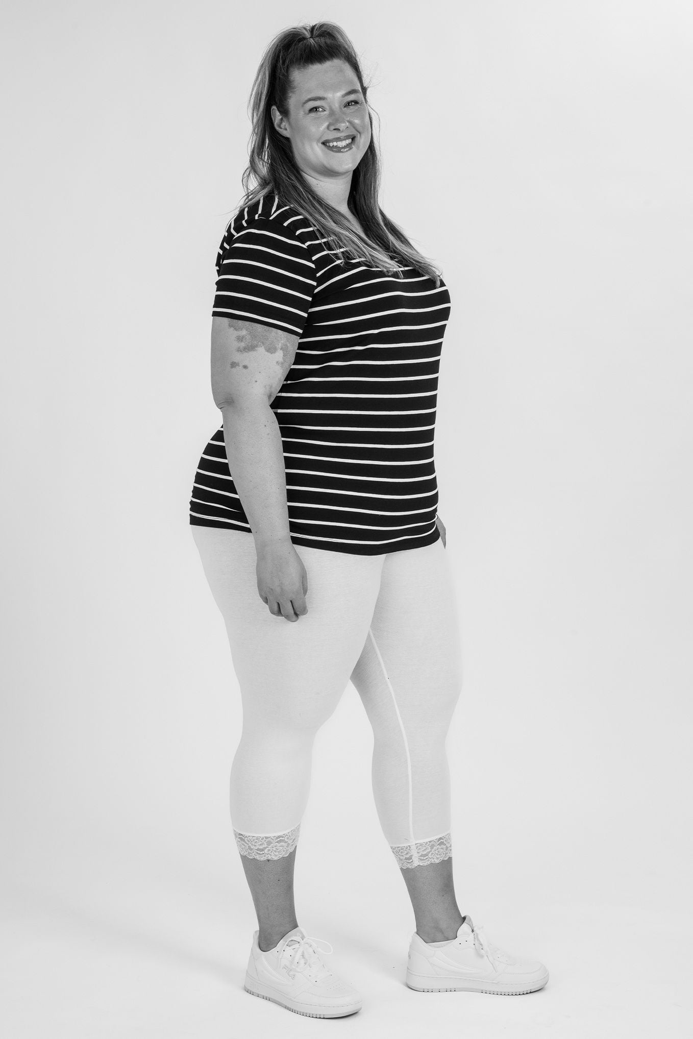 Kvinna i randig t-shirt och vita leggings stående på en vit bakgrund, ler mot kameran. Hennes outfit kombinerar stil och komfort, vilket gör den idealisk för avslappnade aktiviteter eller träning.