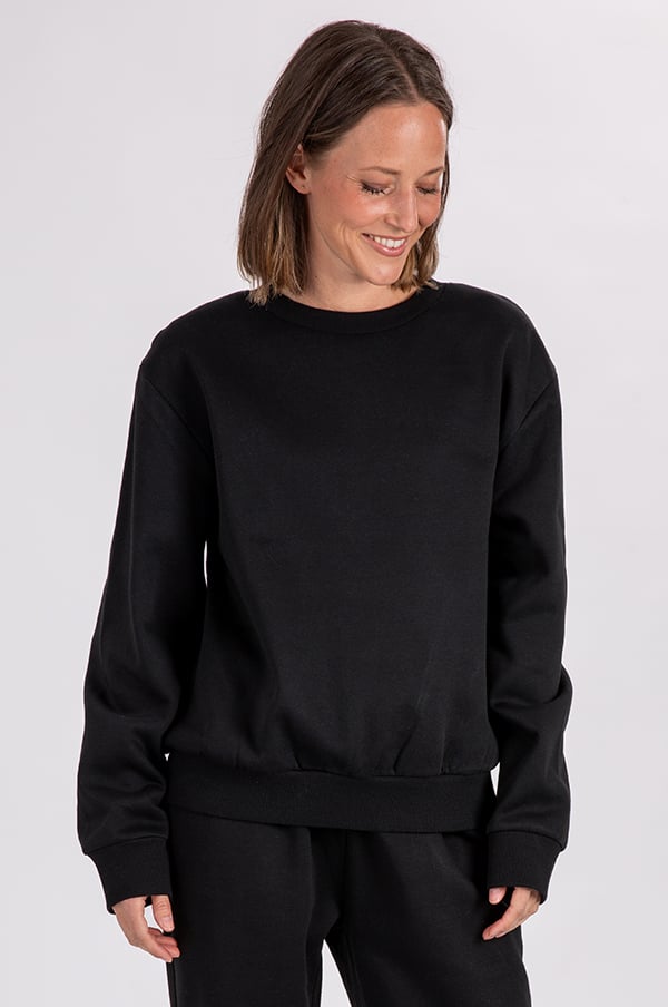 Kvinna i svart sweatshirt med lång ärm och avslappnad passform, ler och ser neråt. Perfekt för både avslappnade och sportiga tillfällen.