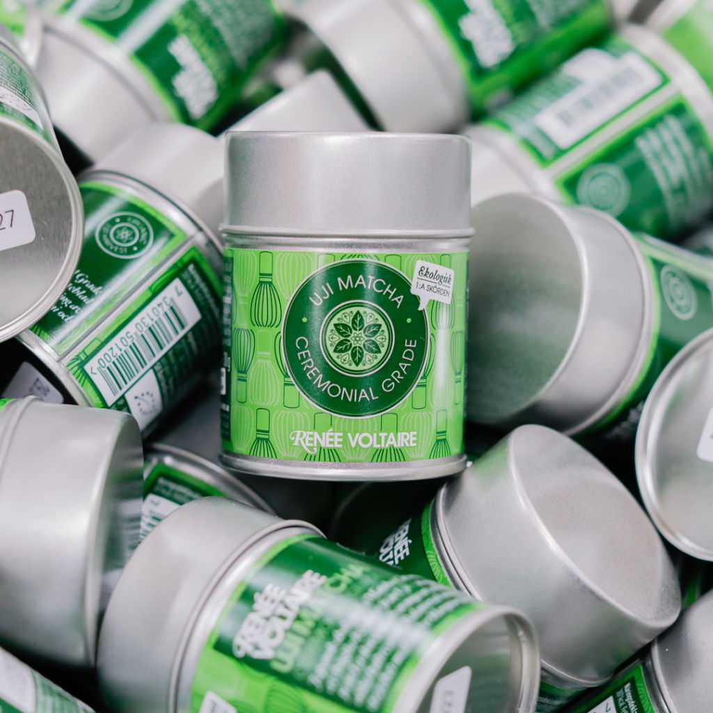 Burkar med Uji Matcha ceremoniell kvalitet från Renee Voltaires sortiment, staplade i en grön och ekologisk miljö.