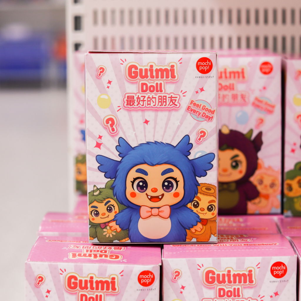 En färgglad förpackning av Guimi Doll med en söt blå karaktär i fokus, omgiven av andra figurer. Produkten är en del av Mochi Pop-serien och betonar teman som vänskap och glädje.