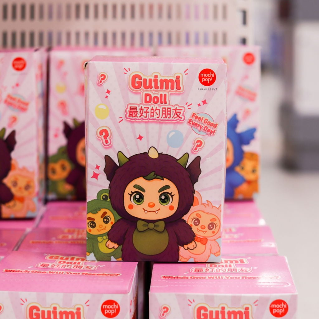 Förpackning av Guimi Doll från Mochi Pop, med en söt karaktär i lila dräkt och färgglada figurer i bakgrunden. Designad för att vara en rolig och överraskande leksak, med text som "Feel Good Every Day!" och frågetecken för att väcka nyfikenhet.