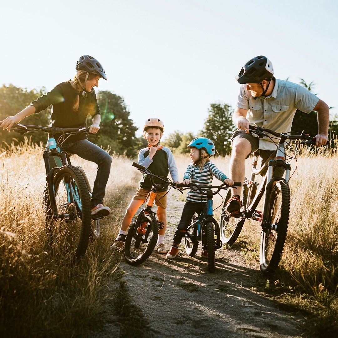 Familj som cyklar på en stig i naturen, med två barn och två vuxna, alla iförda hjälmar. Soligt väder och grönskande omgivningar skapar en glad och aktiv atmosfär.