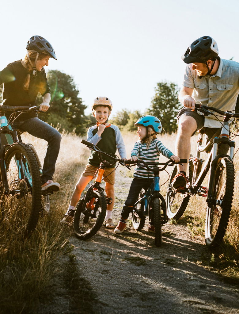 Familj som cyklar tillsammans på en grusväg i naturen. Två vuxna och två barn, alla bär hjälmar, skrattar och interagerar. Soligt väder och omgivande grönska skapar en trevlig atmosfär för utomhusaktiviteter.